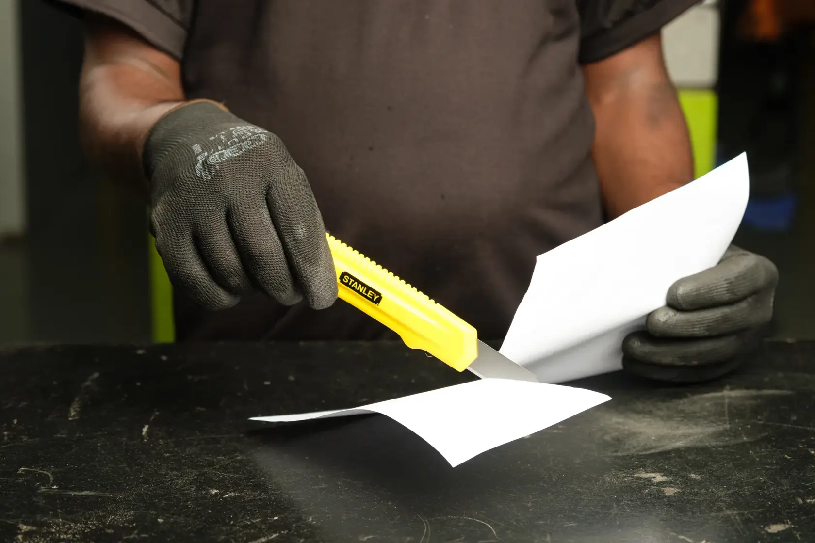 STANLEY 10-143-S yellow utility knife cutting paper, held by gloved hands on a dark table.