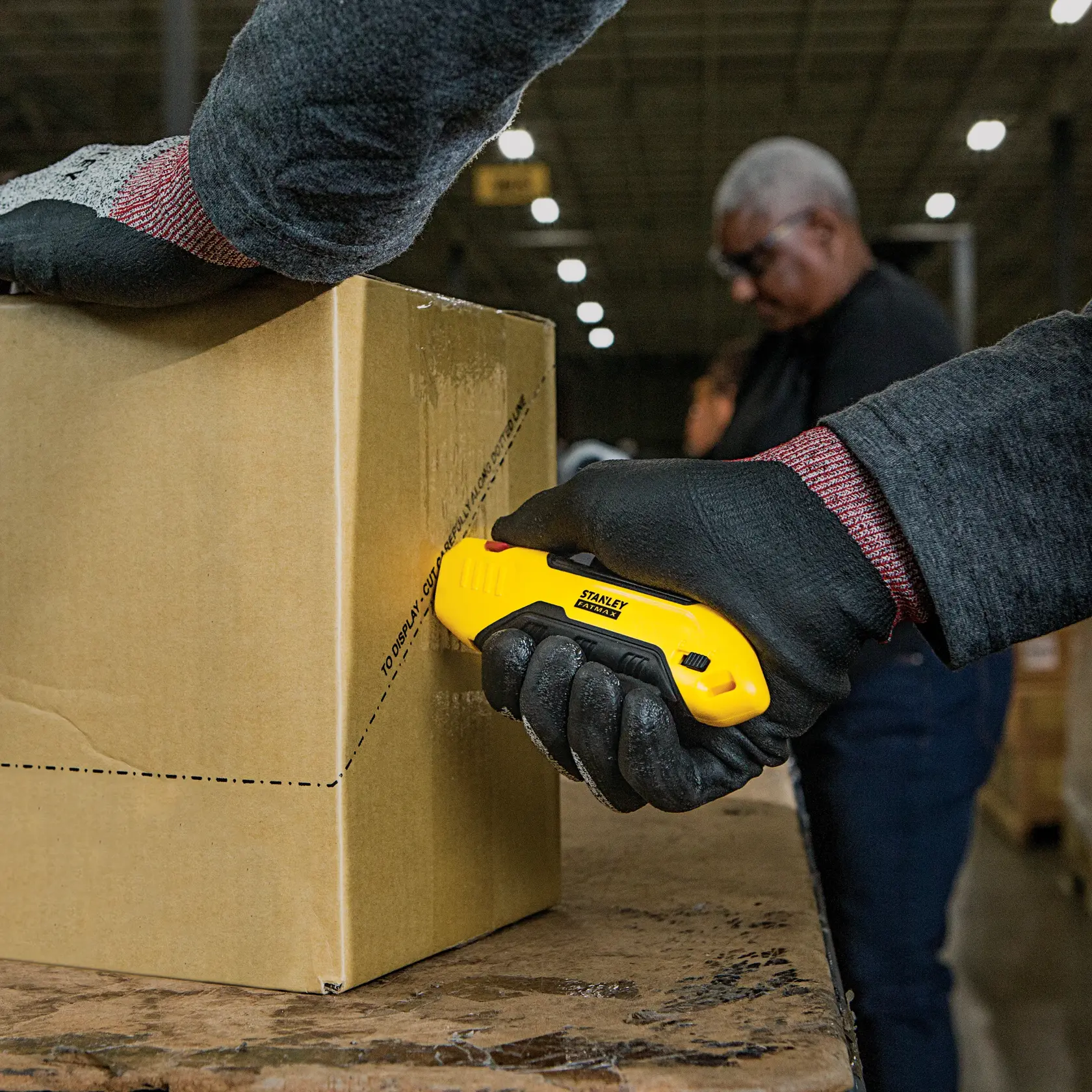 Stanley FMHT10369 utility knife cutting cardboard box, close-up, worker wearing black gloves.