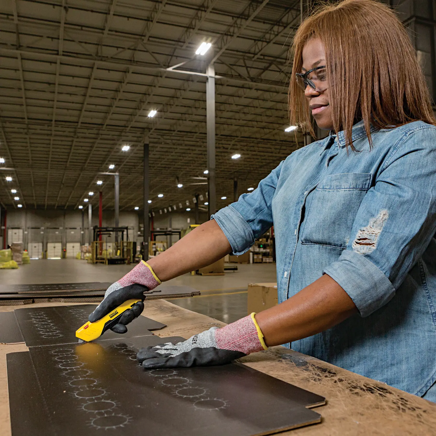 Photo of STANLEY FMHT10369 utility knife cutting black sheet in warehouse by a person wearing safety gloves.