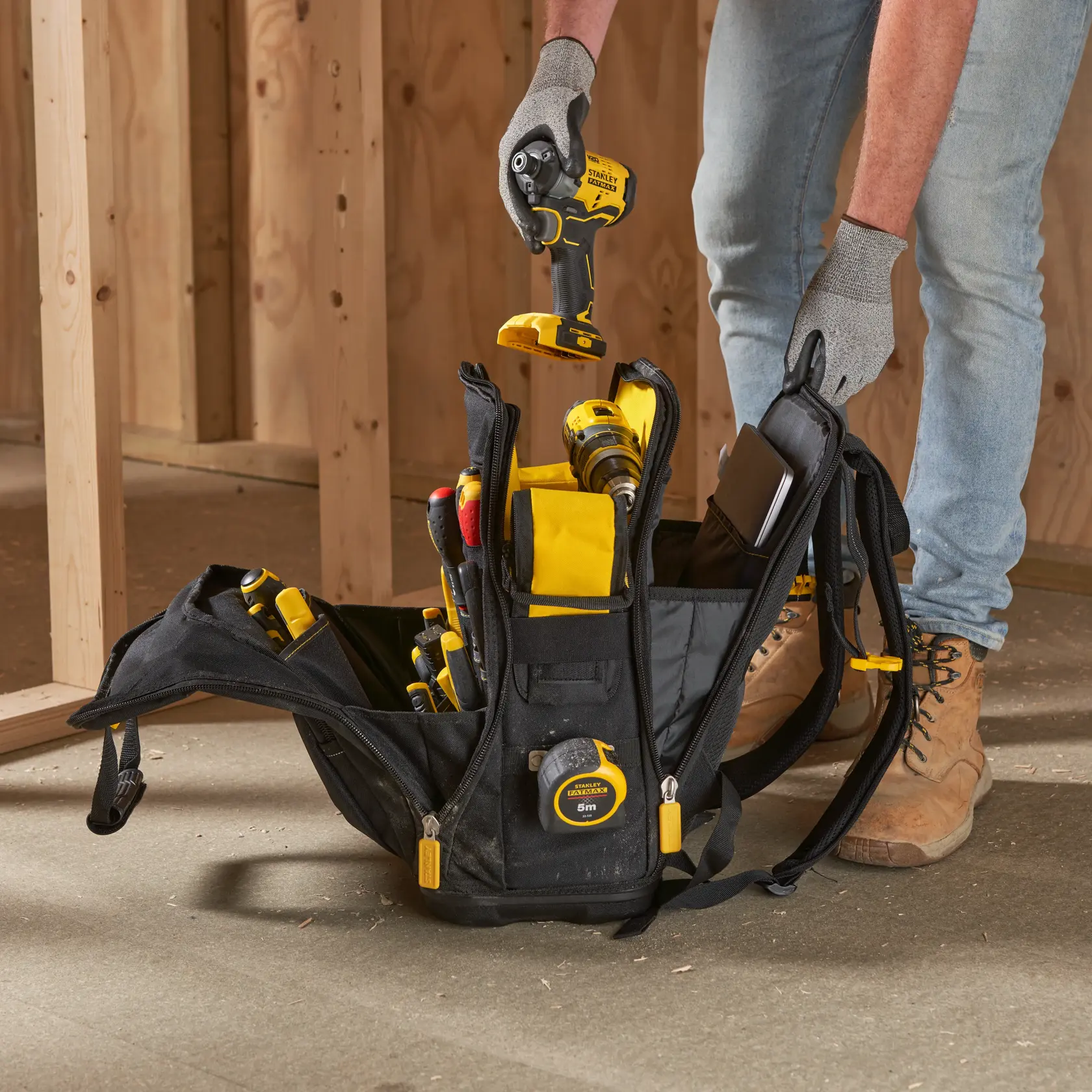 STANLEY FMST1-80144 tool backpack with yellow and black tools on job site floor, worker's hands visible.