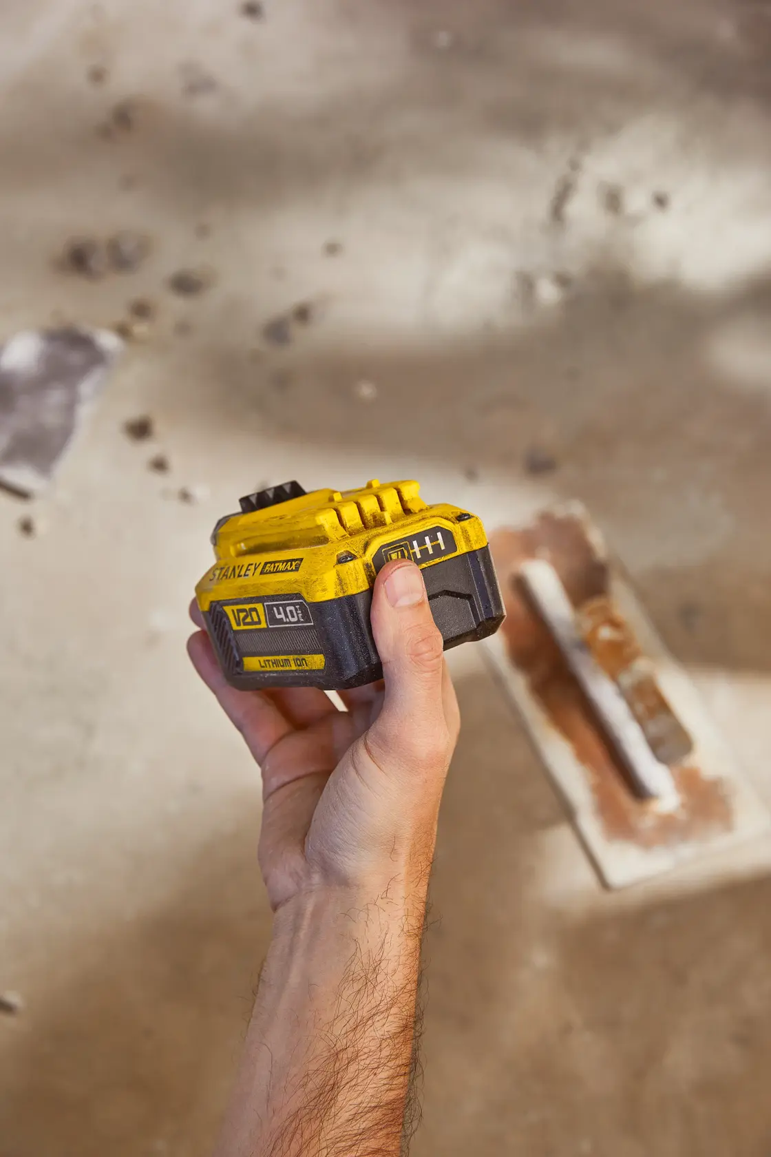 Photo of a STANLEY SB204-KR 20V lithium-ion battery held in hand, with construction tools on ground.
