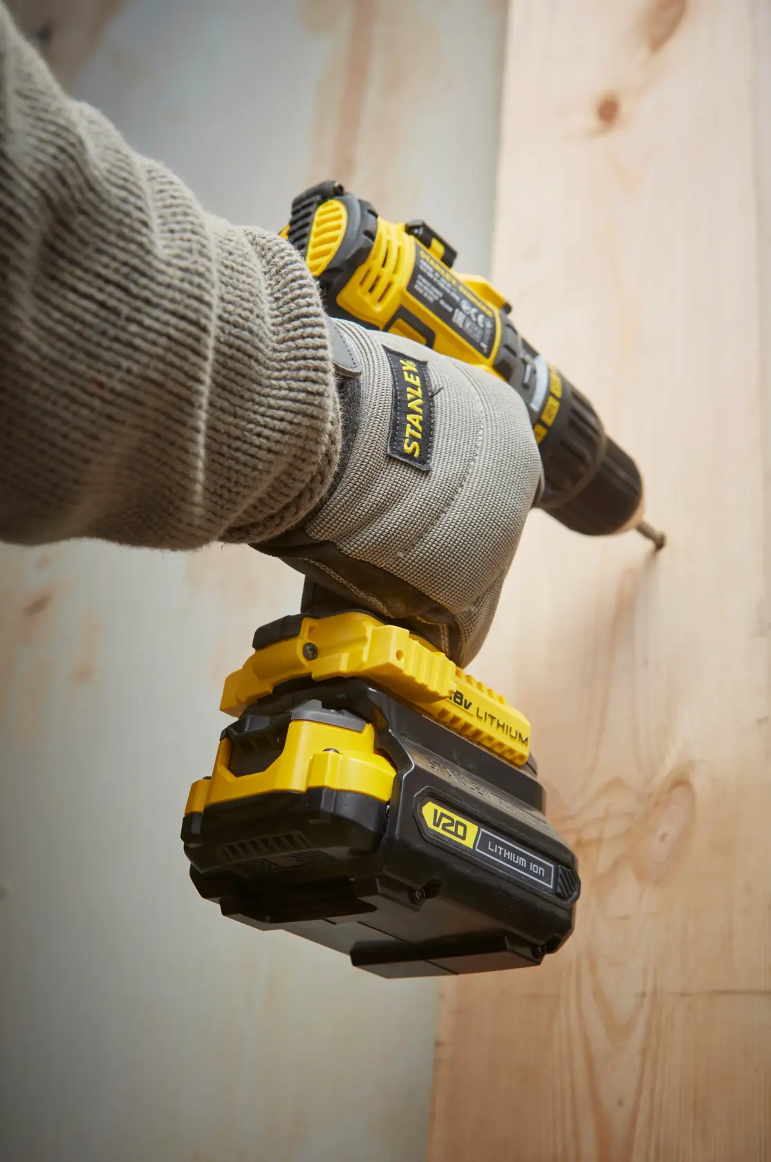 Stanley SBA100 cordless drill held by gloved hand, drilling into wood, side angle, yellow and black tool.