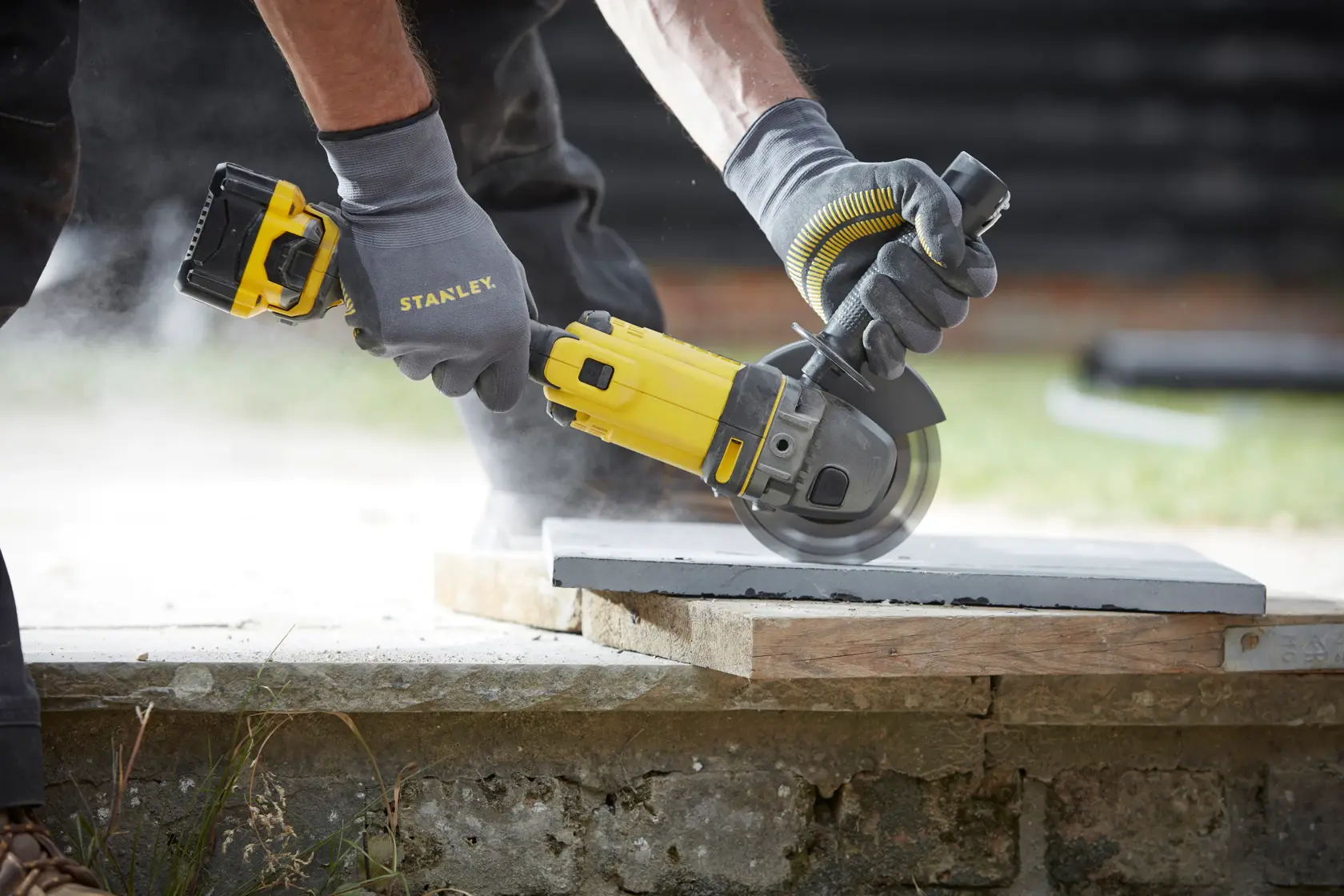 Photo of STANLEY SCG400 cordless angle grinder cutting a concrete slab outdoors, held by gloved hands.