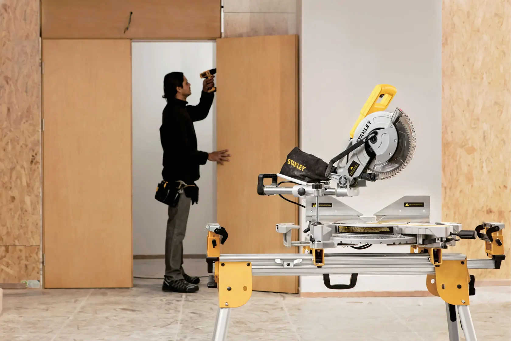 STANLEY SM18-B2C miter saw on stand in workshop, person using drill near wooden door in background.