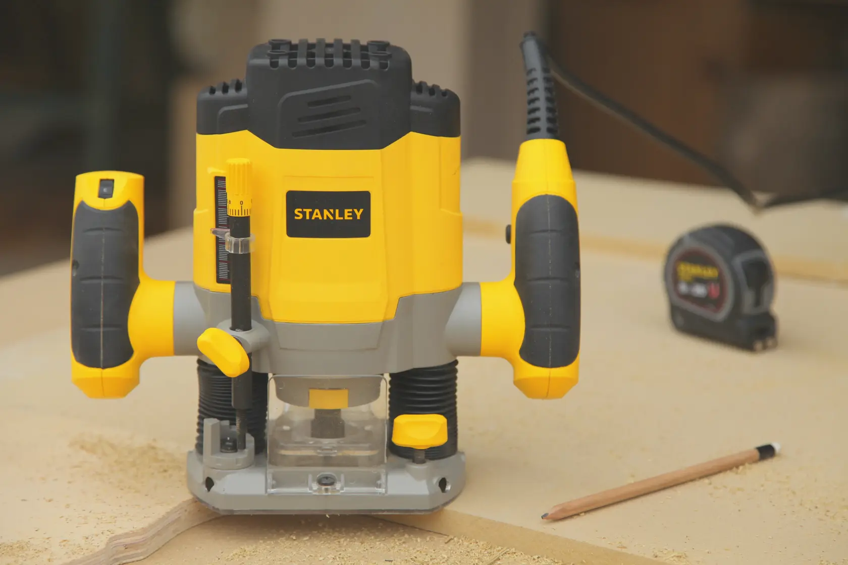 Stanley SRR1200-AR router tool in yellow and black, front angle, placed on a workbench with pencil and tape measure.