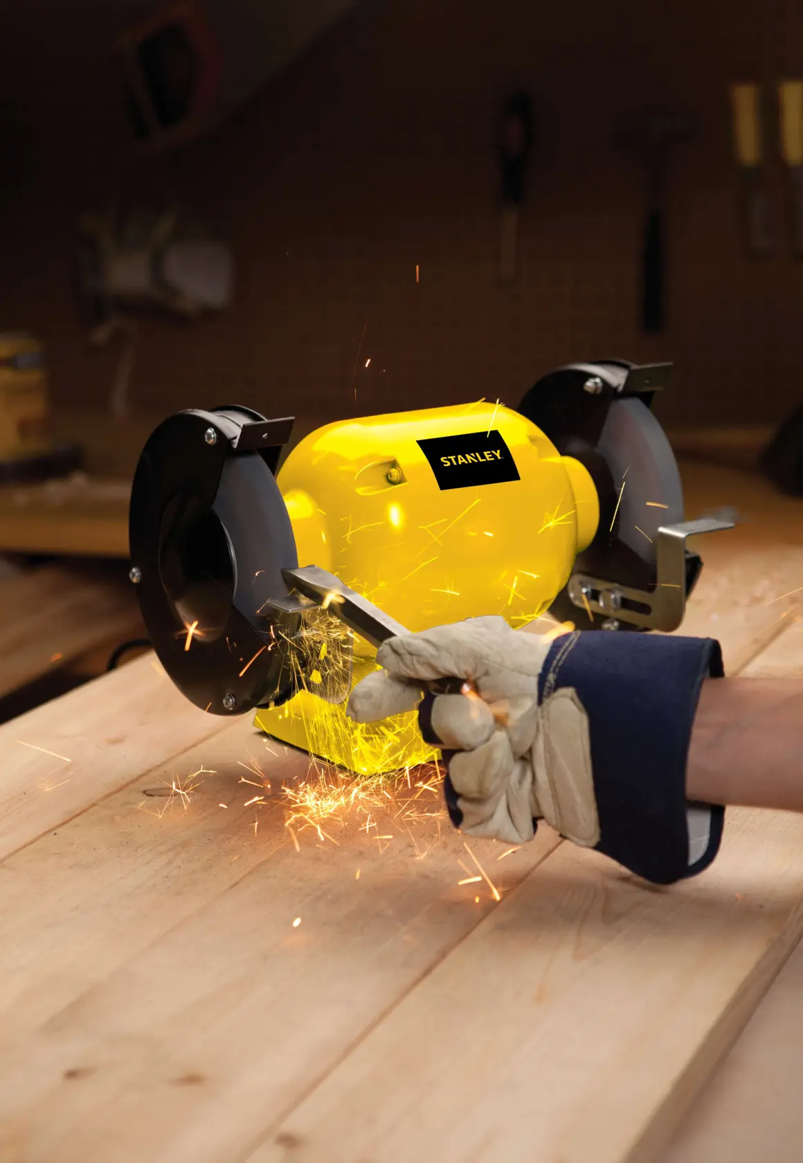 STANLEY STGB3715 bench grinder in use on wooden workbench, sparks flying as gloved hand grinds metal tool.
