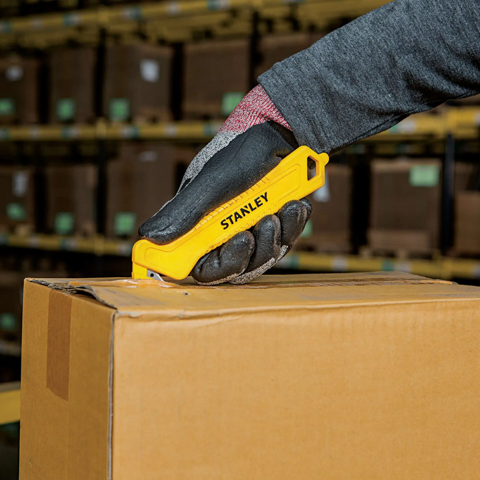 Photo of STANLEY STHT10355 yellow utility knife cutting cardboard box in warehouse setting.