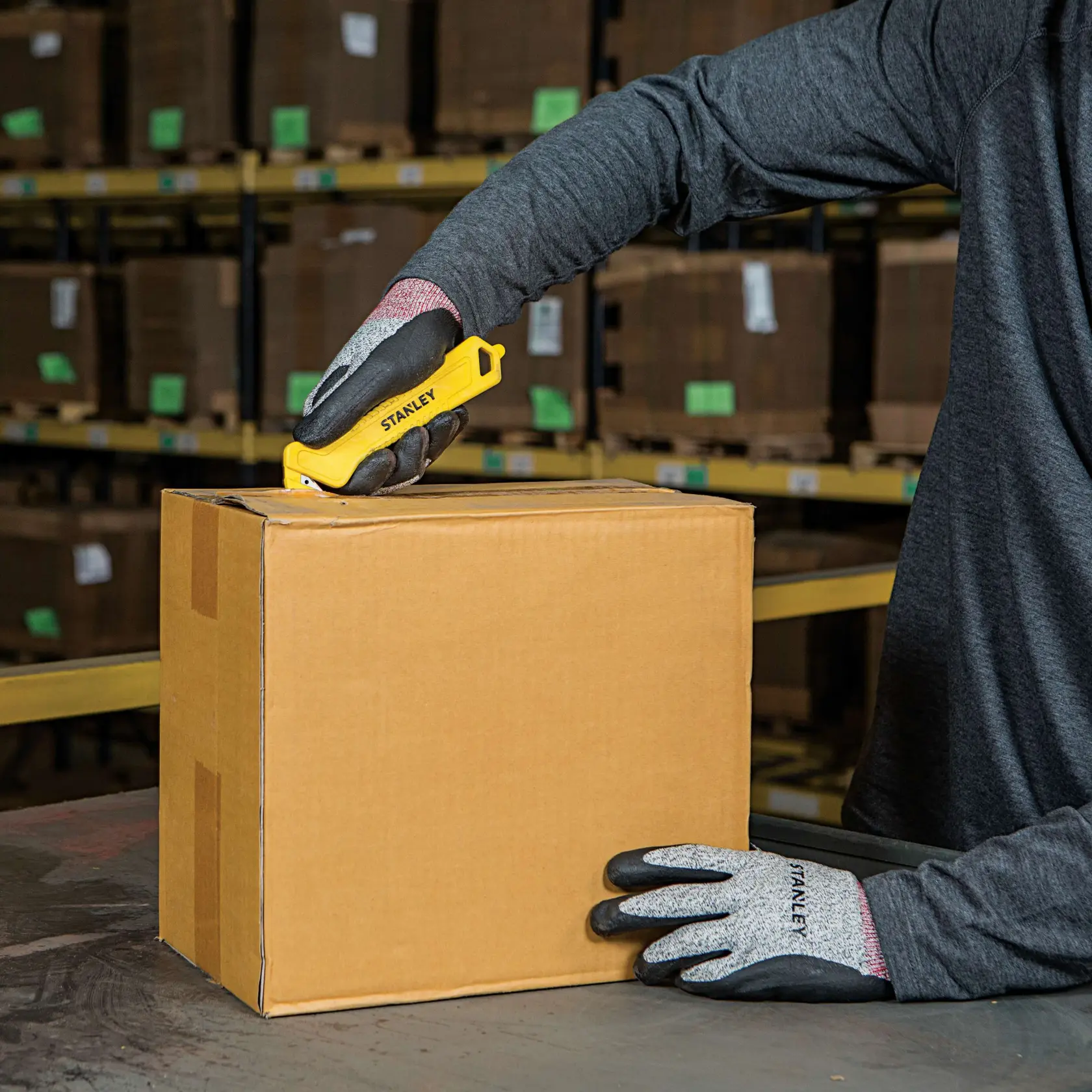 STANLEY STHT10355 utility knife cutting a cardboard box, worker wearing gloves, warehouse background.