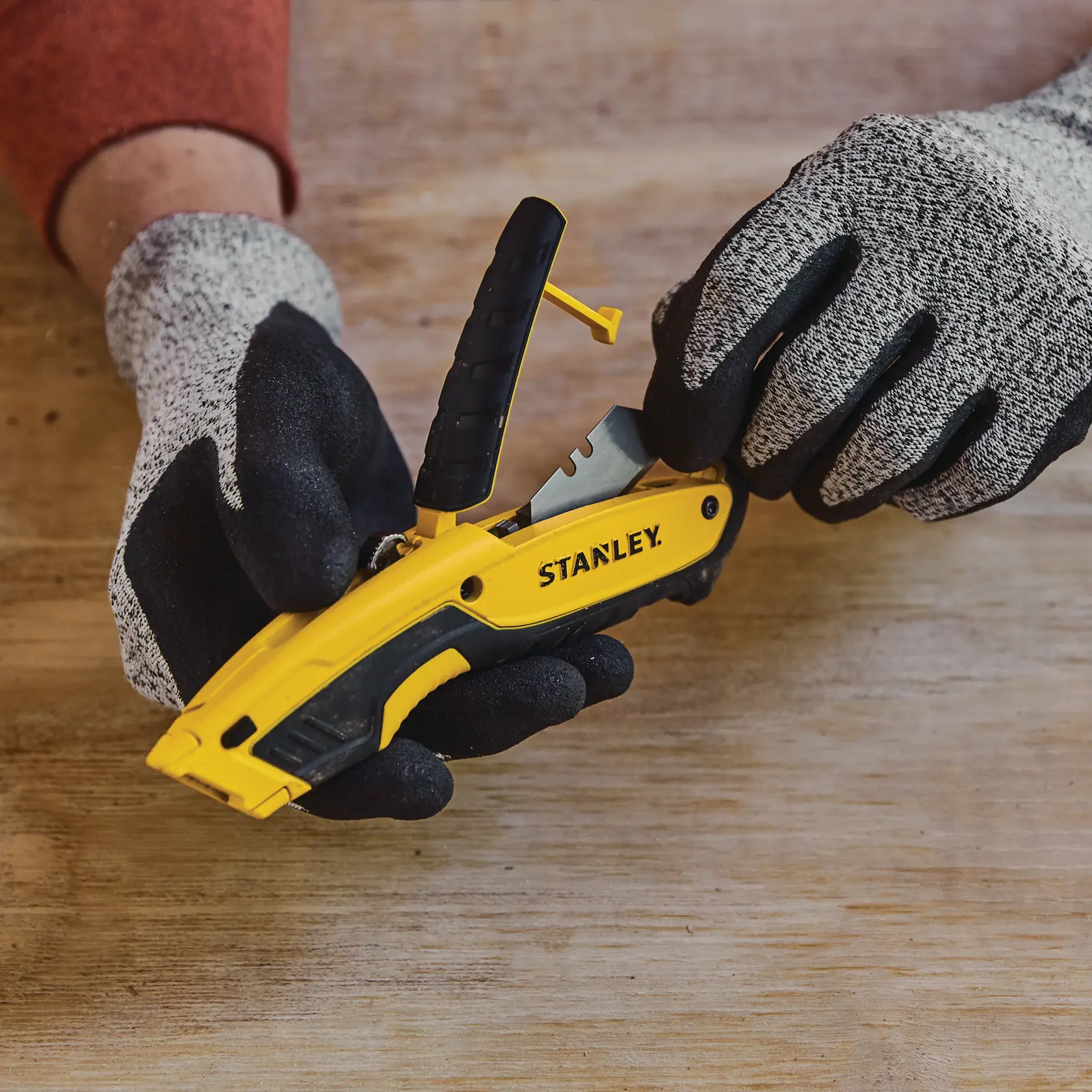 Photo of STANLEY STHT10479-0 yellow utility knife with black handle held by gloved hands on wooden surface.